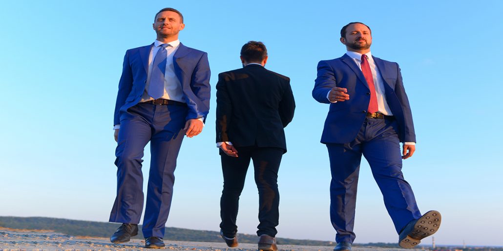 Three men dressed in suits walking accross open ground. Two facing front and one facing back.